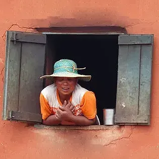 Femme souriante en chapeau de paille penchée à une fenêtre en bois rustique à Madagascar